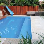 A modern pool with outdoor furniture and a water slide next to a glass wall